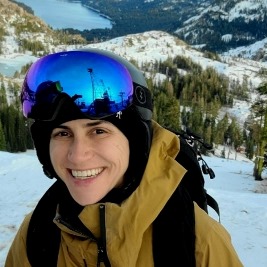 Lee Morris smiles at the camera, wearing her ski goggles and a yellow winter coat in front mountains covered with snow and evergreen trees.