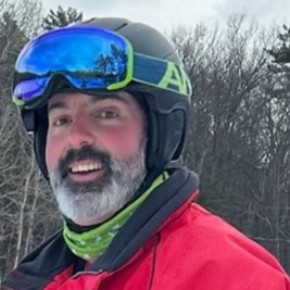 Mike Reilly smiles at the camera, wearing his ski goggles and a red winter coat in front of a forest in winter.